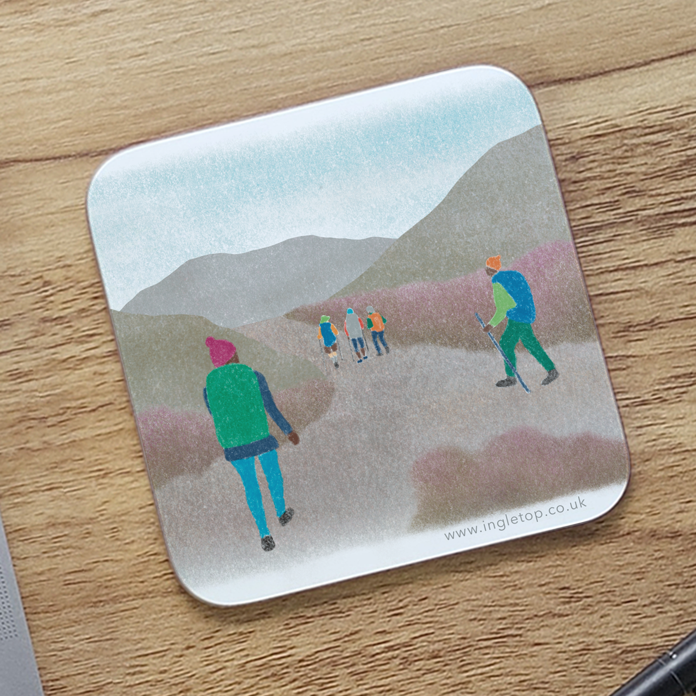 A square coaster with rounded corners, printed with an illustration of hikers heading into the hills, lying on a wooden desk