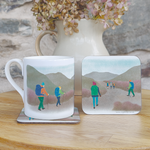 A fine china mug sitting on a hardboard coaster, with another coaster standing beside them. They are decorated with an illustration of a group of hikers heading in to a hilly landscape