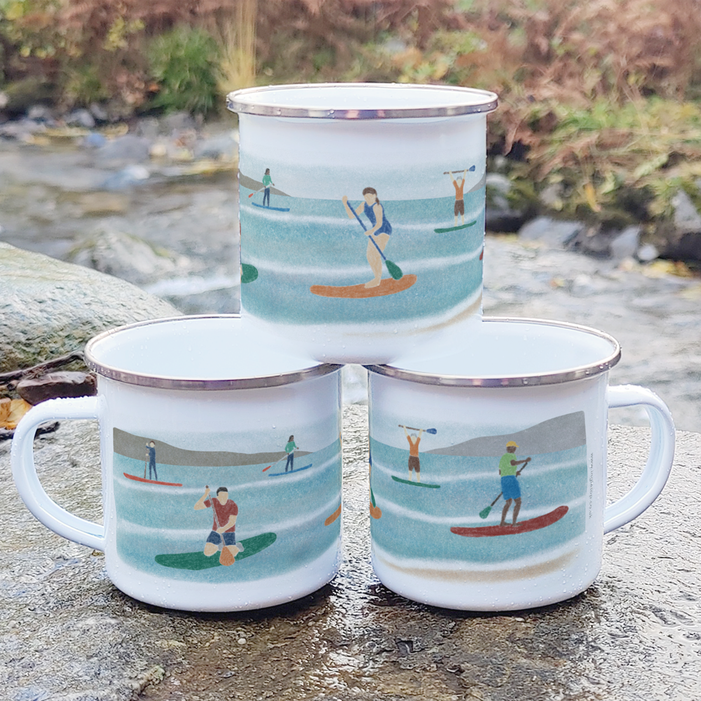 Three stacked enamel mugs, printed with an illustration of a group of six paddle boarders, displayed outdoors on a rock