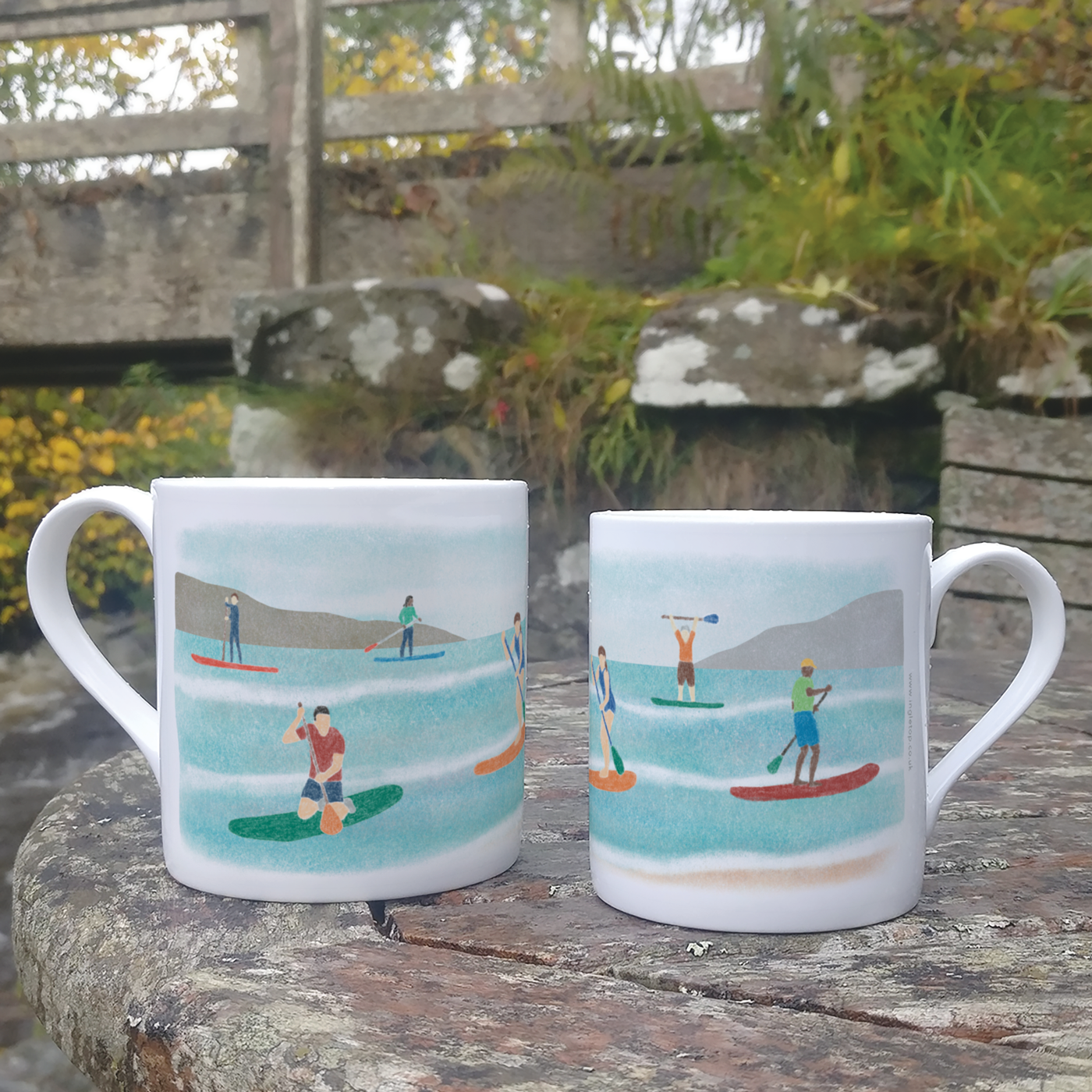 Two different sized mugs, decorated with an illustration of paddleboarders, on an outdoor table