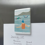 A rectangular ceramic magnet, holding a shopping list on to a fridge, decorated with an illustration of three wild swimmers in the water